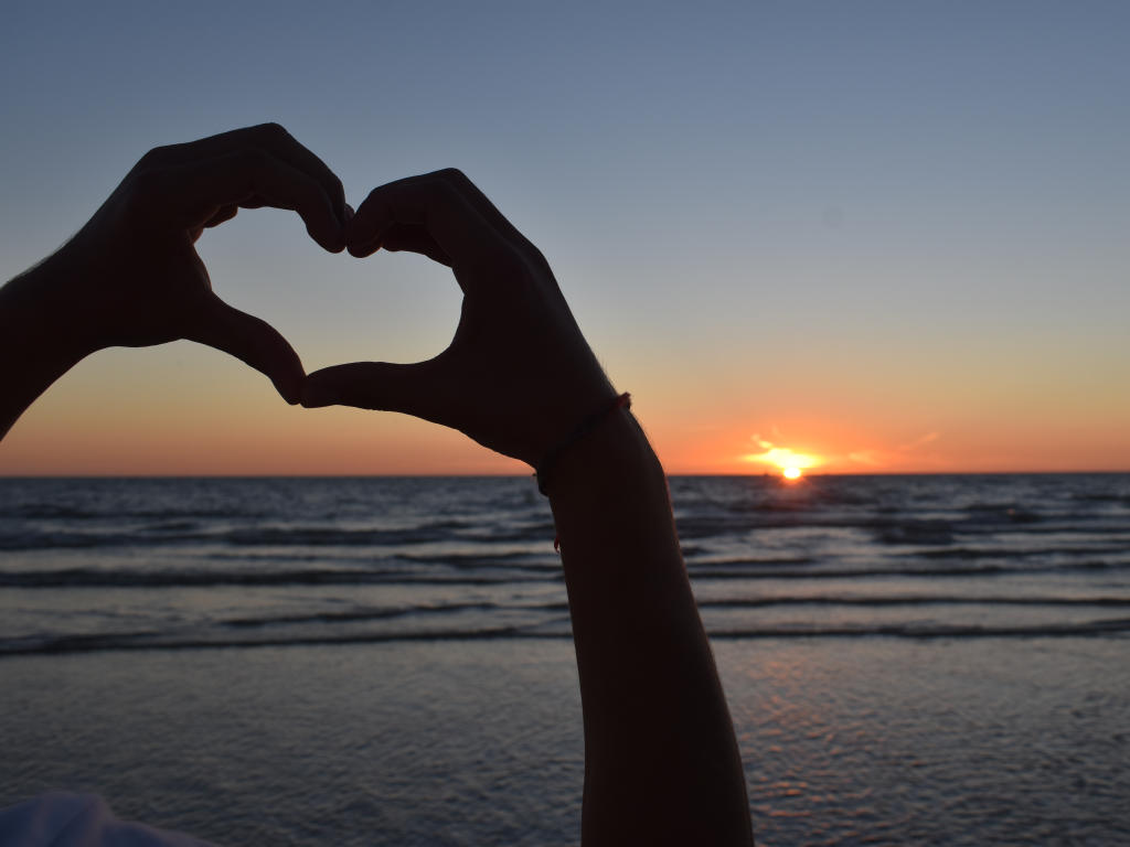 Hände welche ein Herz formen mit dem Meer und dem Sonnenuntergang als Hintergrund